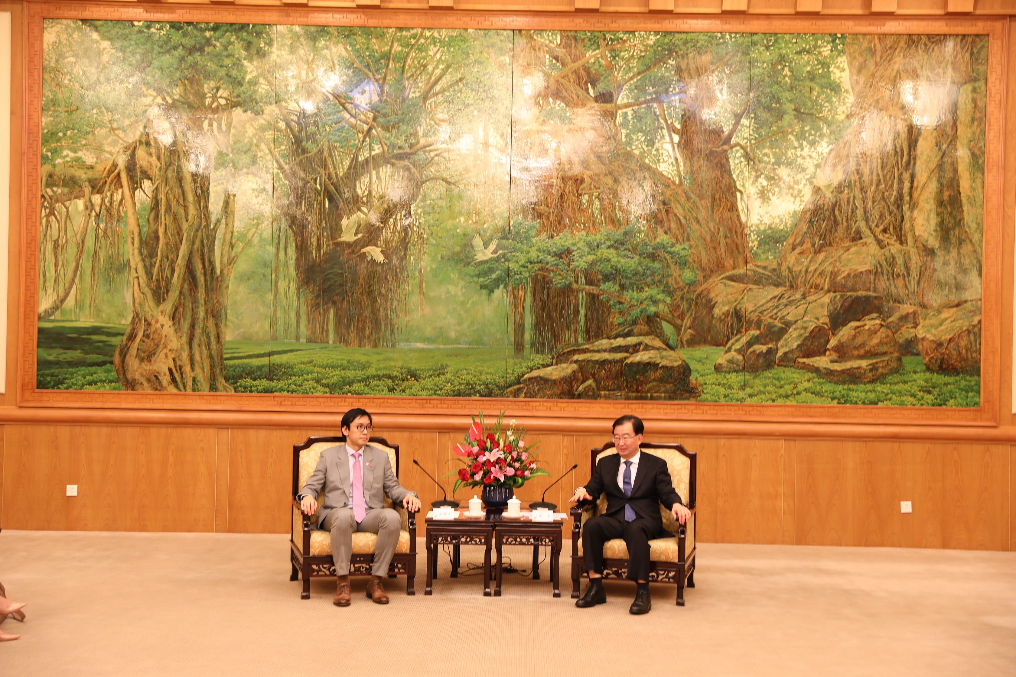 Two men in suits seated below large tree painting in wood-paneled room.